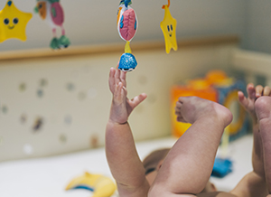 A baby girl in a diaper lying in a cot reaching up to a colourful mobile hanging above her.