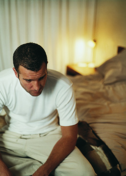 A man in pajamas is sitting on the side of a messy bed. He looks tired.