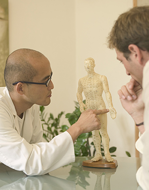 Male acupuncturist by patient, pointing at acupuncture point on model.