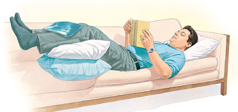 Man lying on couch, reading, with legs supported by pillows. Ice pack is on front of lower legs.