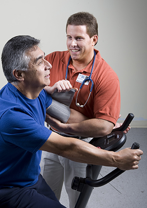 Man sitting on an exercise bike. A physical therapist is standing next to her and watching.