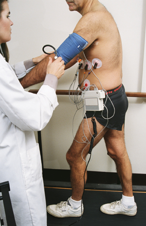 Man with wires attached to chest walking on treadmill. Wires go to ECG machine next to treadmill. Healthcare provider is monitoring ECG machine.