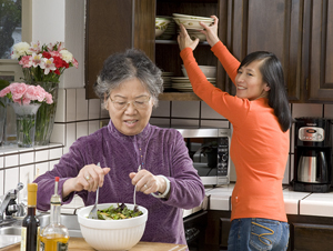 Older woman preparing salad in kitchen, younger woman reaching up into cupboard for bowls.