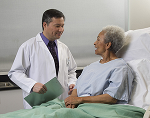 Senior woman wearing a hospital gown in hospital bed talking with male Doctor.