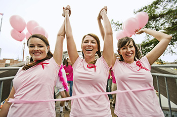 Three women wearing pink ribbons are holding hands in the air and celebrating