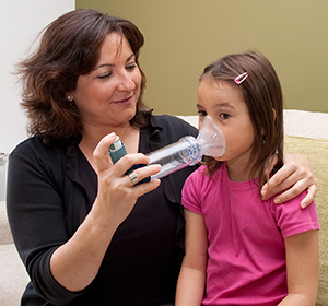 Woman helping girl use mask inhaler with spacer.