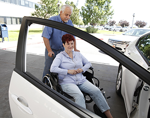 Woman in wheelchair, partner helping her into car in parking lot.