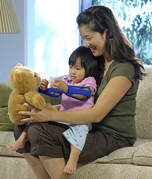 Toddler with arm restraints playing with teddy bear while sitting in woman's lap.